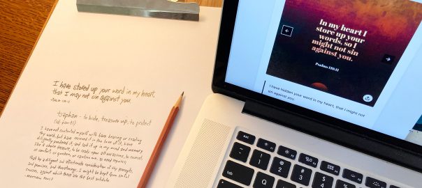 Bible verse written on paper with pencil, sitting next to a laptop with the same Bible verse